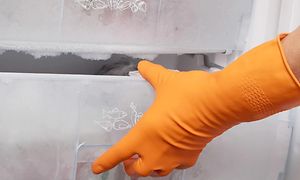 Hand i orange diskhandske öppnar en igenfrusen fryslåda i en frys som behöver frostas av.