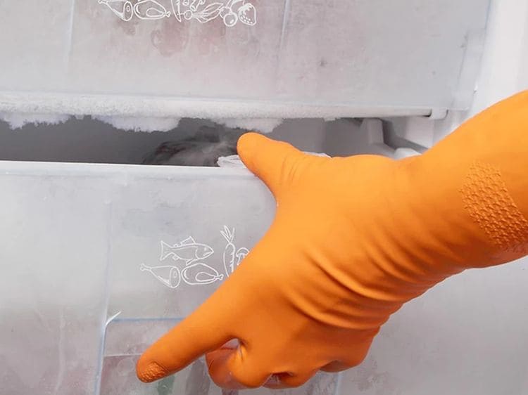 Hand i orange diskhandske öppnar en igenfrusen fryslåda i en frys som behöver frostas av. 