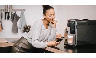En kvinna står och ser på en mobil framför en Melitta kaffemaskin med en färdigbryggd kaffe latte. 