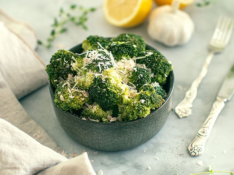 En skål med broccoli med lite ost på toppen på ett köksbord med bestick, en citron och en vitlök vid sidan. 