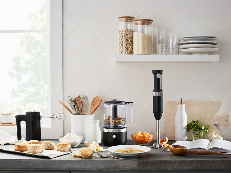 Matberedare, stavmixer och blenders uppställda på en köksbänk bredvis olika slags matvaror.