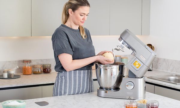 Kvinna med förkläde i ett kök lyfter upp deg från skålen i en Kenwood köksmaskin 