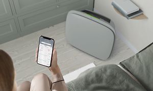 En grå Well 5 på golvet och en kvinna med Wellbeing-appen på sin telefon