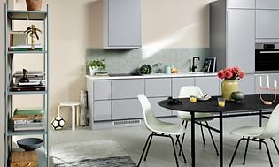 Ljusgrått EPOQ Integra kök med öppen planlösning, ett svart köksbord med vita stolar och en bokhylla. 