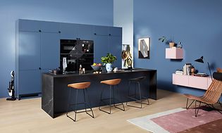 Blått EPOQ Trend kök med köksö i mitten med barstolar. Väggarna är också blå med rosa hyllor. Integrerad ugn och kaffemaskin.