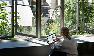 Kvinna som sitter vid ett skrivbord bredvid ett fönster och arbetar på hennes bärbara dator