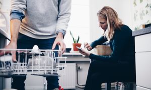 En man står vid en öppen diskmaskin med disk i och en kvinna sitter vid bordet med sin telefon