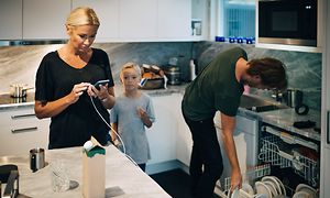 Familj i kök, kvinna med sin telefon med ett barn bredvid sig, mannen står framför en öppen diskmaskin