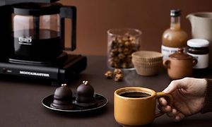 SDA - Kaffebryggare - Hand som håller en kopp framför efterrätt och Moccamaster kaffebryggare på ett bord