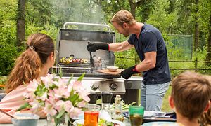 Man som grillar mat på en gasolgrill medan hans familj tittar på