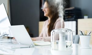 En luftfuktare i ett kontor med en kvinna som ler och sitter vid ett skrivbord i bakgrunden