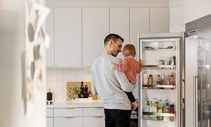 Pappa som håller sitt barn i ett kök bredvid ett öppet kylskåp