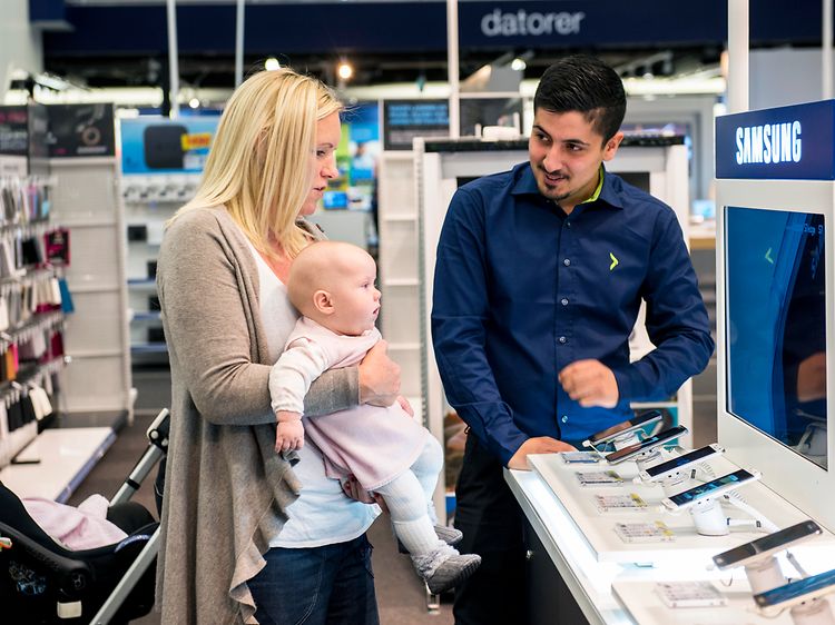 Mamma med baby i famnen får hjälp av en butiksanställd i en Elgiganten-butik. De står vid ett Samsung bord med telefoner.