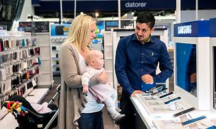Mamma med baby i famnen får hjälp av en butiksanställd i en Elgiganten-butik. De står vid ett Samsung bord med telefoner.