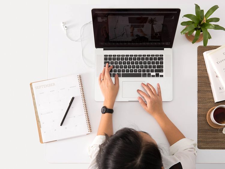 Bild ovanifrån på ett vitt bord med en macbook på och person som sitter och jobbar. En kaffekopp, böcker och krukväxt vid sidan.