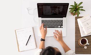 Bild ovanifrån på ett vitt bord med en macbook på och person som sitter och jobbar. En kaffekopp, böcker och krukväxt vid sidan.
