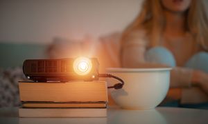 Kvinna sitter i soffan och ser på en film som projekteras via en hemmabio-projektor som är uppställd på en hög med böcker.