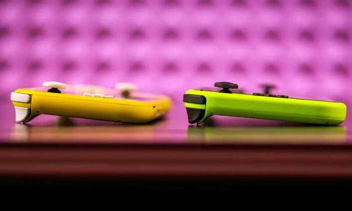 Nintendo Switch Lite, två spelkonsoler på ett bord, en i beige färg och den andra grön. Rosa vägg i bakgrunden.