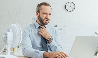 En man som är för varm och sitter framför datorn med en fläkt på skrivbordet