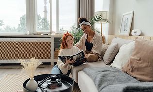 Två kvinnor i en soffa, läser en bok och har en luftkvalitetsmätare bakom sig