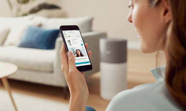 Kvinna sitter i ett vardagsrum med Philips air luftrenare bredvid sig och Clean Home+ app på sin smartphone
