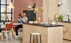 Man sitter och håller i en baby vid ett köksbord i ett kök från Epoq i ljus träfärg med köksö i mitten. 