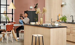 Man sitter och håller i en baby vid ett köksbord i ett kök från Epoq i ljus träfärg med köksö i mitten.
