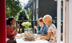 Tre glada kvinnor sitter runt ett bord och äter på en solig balkong 