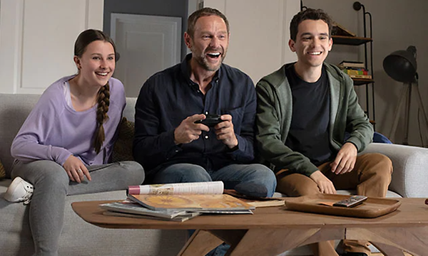 Tre personer sitter i en grå soffa, en vuxen man och två ungdomar. Mannen i mitten håller i en Nintendo Switch-enhet. 