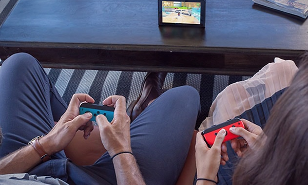 Bil ovanifrån på två personer som spelar Nintendo Switch med joycon-kontroller i händerna och bildskärm på bordet framför.  