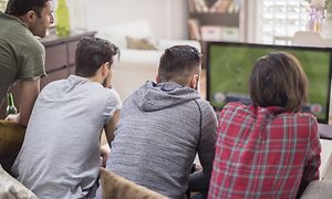 Fyra män sitter tätt ihop i en soffa och tittar på fotboll på TV.