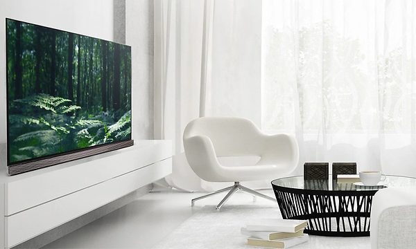 En stor OLED LG TV i ett ljust vardagsrum helt i vitt. TV'n står på en vit tvbänk med en fåtölj och ett litet soffbord i glas. 