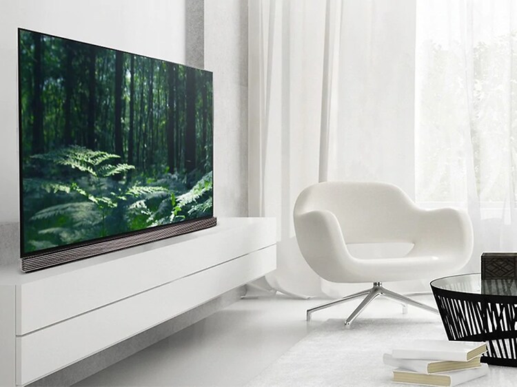 En stor OLED LG TV i ett ljust vardagsrum helt i vitt. TV'n står på en vit tvbänk med en fåtölj och ett litet soffbord i glas. 