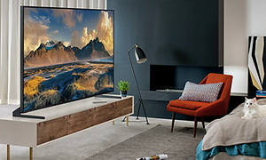 En väldigt stor TV på en tv-bänk i ett modernt rum. En vit katt sitter på en säng och en röd fåtölj är placerad framför TV:n.