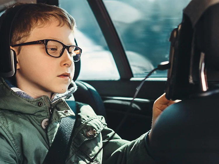 Pojke i baksätet i bilen sitter med hörlurar på och tittar på en surfplatta som är fäst på nackskyddet på förarsätet.