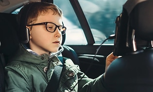 Pojke i baksätet i bilen sitter med hörlurar på och tittar på en surfplatta som är fäst på nackskyddet på förarsätet.