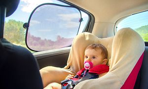 En liten bebis sitter i bilbarnstol i baksätet, och ser framåt med en rosa napp i munnen. Ett solskydd är fäst på fönsterrutan. 