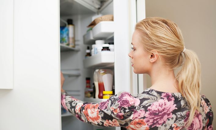 En blond kvinna i blommig tröja öppnar kylskåpet och sträcker sig efter något.