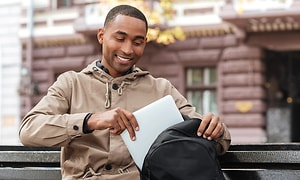 En man i beige jacka sitter på en parkbänk och packar ner sin laptop i en svart ryggsäck. 