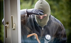 En inbrottstjuv med rånarluva tittar in genom ett fönster hållandes i en kofot