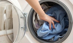 Tvättmaskin med luckan öppen och en arm som stoppar in en blå handduk i maskinen som redan innehåller en del tvätt. 