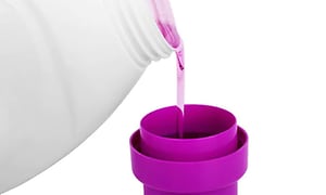 Flytande tvättmedel i rosa färg hälls upp från en vit flaska i ett lila lock. 