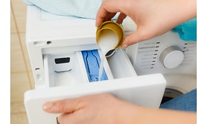 Närbild på hand som fyller flytande tvättmedel i tvättmaskinen, med hjälp av toppen på flaskan. 
