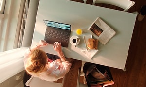 Bild ovanifrån på en kvinna som jobbar på en bärbar dator vid ett skrivbord med en macka, juice och kaffe vid sidan av. 