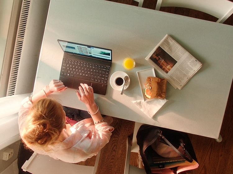 Bild ovanifrån på en kvinna som jobbar på en bärbar dator vid ett skrivbord med en macka, juice och kaffe vid sidan av. 