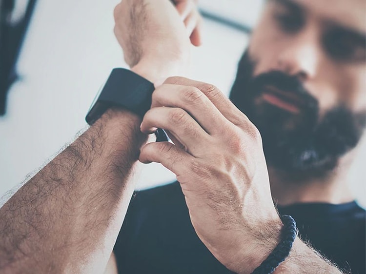 En man med skägg tar på sig en svart smartklocka och spänner åt runt handleden. 