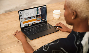 En kvinna med kort blont krulligt hår sitter framför en Lenovo yoga slim dator, en take away kopp står brevid på bordet.