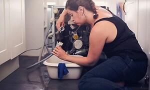 Kvinna som sitter vid baksidan av en tvättmaskin och håller upp en slang som hon tittar skeptiskt på. 