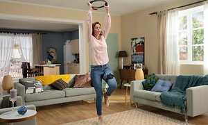 En kvinna tränar i vardagsrummet med Nintendo Ring fit, hon har en ring upplyft ovanför huvudet och ett band runt låret. 