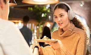 Kvinna med ljusbeiga on-ear hörlurar på sig håller fram en tallrik med mat på en restaurang. 
